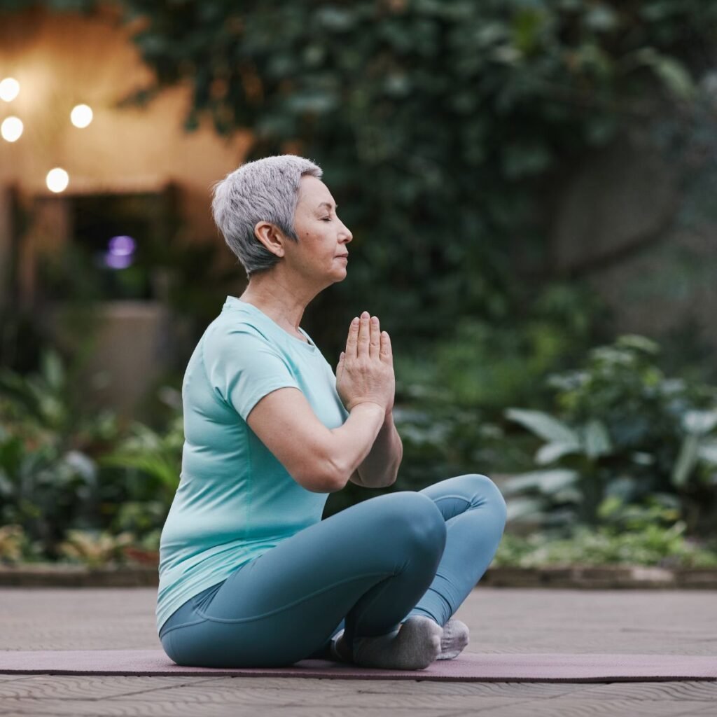 Outlive também aponta como pilar de uma vida longa a prática da meditação e outras técnicas que possibilitem a diminuição dos níveis de stress. A imagem mostra uma mulher de meia idade sentada em um jardim meditando e com uma expressão facil que demonstra tranquilidade.