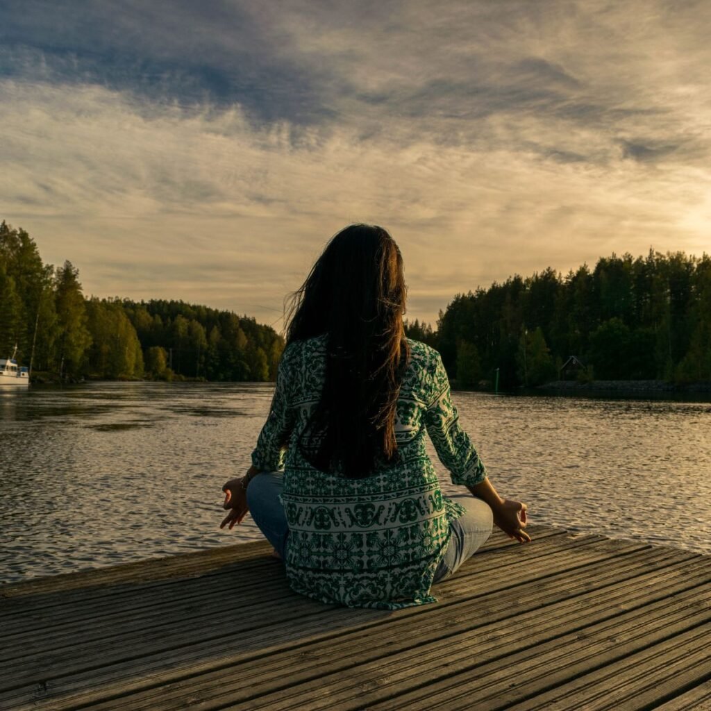 Quanto tempo leva para sentirmos os efeitos da meditação na saúde mental. A imagem mostra uma mulher sentada em posição de lótus em um deque diante de um rio.