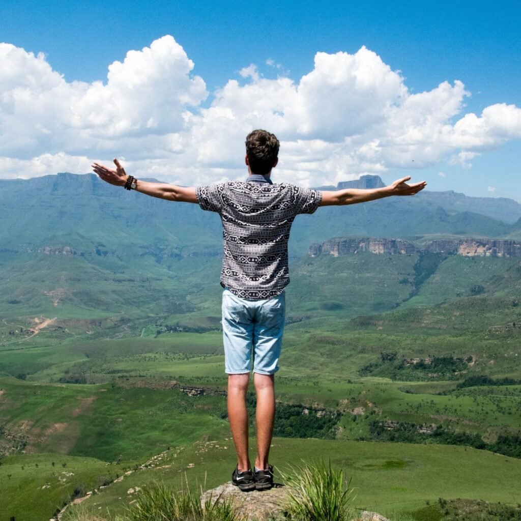 Homem diante do céu e da paisagem verde. Ele está num alto monte.