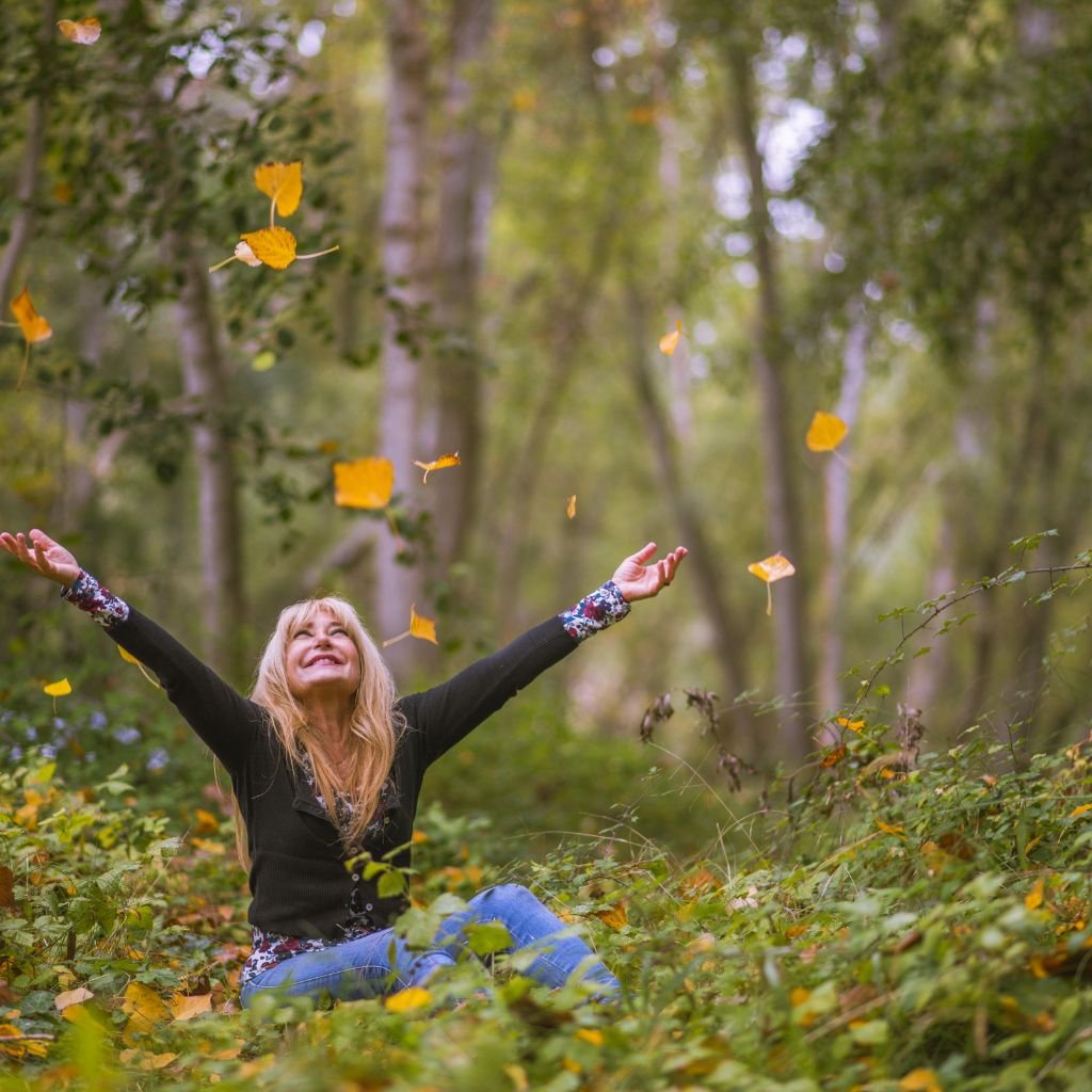 Mulher sentada na relva, jogando folhas para o alto e demonstrando felicidade.